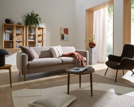Ljus skandinavisk vardagsrumsscen med beige soffa, runt soffbord med tidning och litet ljus, neutral matta och roströd pläd; trähyllor med böcker, växt, abstrakt tavla och stort fönster med skira gardiner i dagsljus.