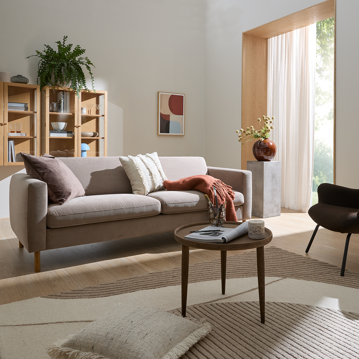 Ljus vardagsrumsinredning med beige soffa, kuddar och pläd, runt soffbord med tidning och ljus, stor matta, öppna hyllor, växt och vas vid ett fönster med skira gardiner.