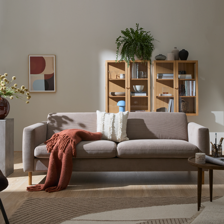 Ljus vardagsrumsinteriör med beige soffa, vit kudde och roströd pläd, väggmonterad bokhylla i trä med växter och keramik, abstrakt väggkonst och mjuk mönstrad matta i dagsljus.