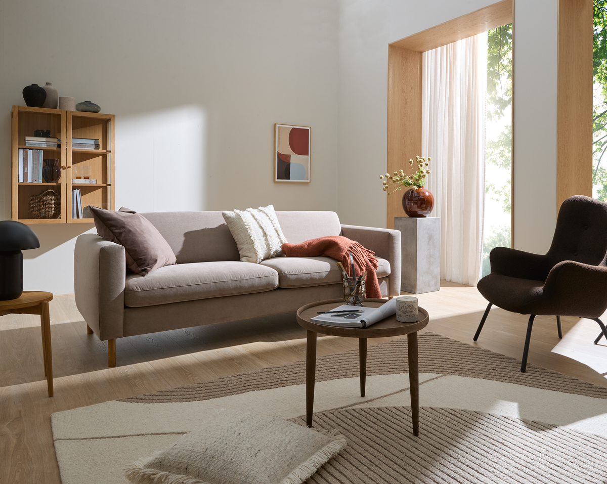 Ljust vardagsrum i neutrala toner med beige soffa, brun fåtölj, runt träbord med tidning och ljus, vägg-hylla och konst, stor fönsternisch med skira gardiner och vas med blommor.