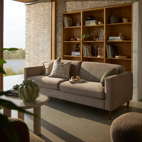 Ljus vardagsrumsbild med beige soffa framför en träbokhylla, soffbord med grön vas och boken Homes for Our Time, sjöutsikt genom stort fönster.