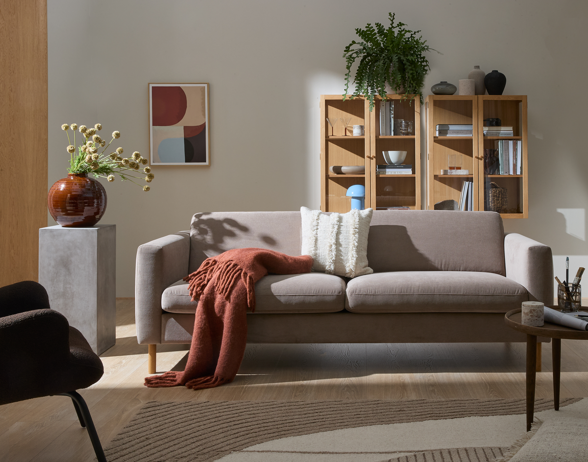 Ljust vardagsrum med beige soffa, rostfärgad pläd och kuddar, väggmonterat trädskåp med böcker, dekorationer, grön växt och liten blå bordslampa; abstrakt tavla, rundt soffbord och vas på piedestal på mönstrad matta i dagsljus.