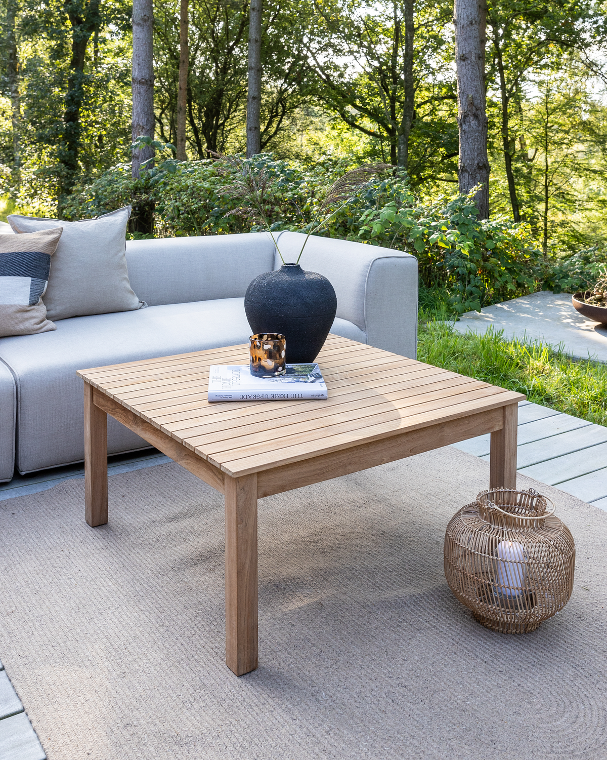 Utomhuslounge med teak soffbord på matta, grå soffa, svart vas med gräs, bok och liten ljuslykta på bordet samt rottinglykta med ljus bredvid, omgiven av grönska.