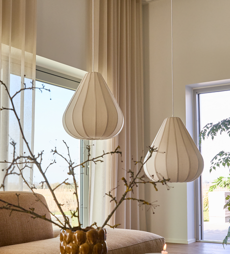 Två beige tygpendlar över soffa vid stora fönster, skira gardiner, vas med kvistar och dagsljus.