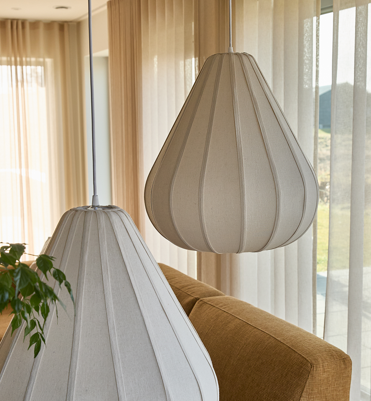 Närbild på två beige textilpendlar ovanför en senapsgul soffa i ett ljust vardagsrum med skira gardiner.