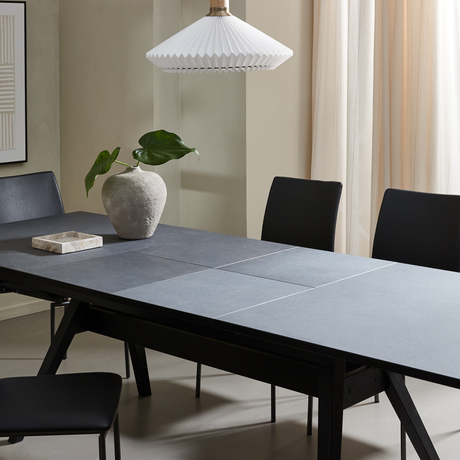 Minimalistisk matplats med mörkt rektangulärt bord, svarta stolar, grå stenvase med gröna blad och vit plisserad pendellampa framför ljusa gardiner.
