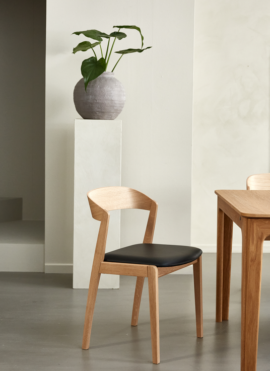 Trästol med svart sits bredvid ett matbord; rund vas med grön växt på piedestal i en ljus, minimalistisk matsal.