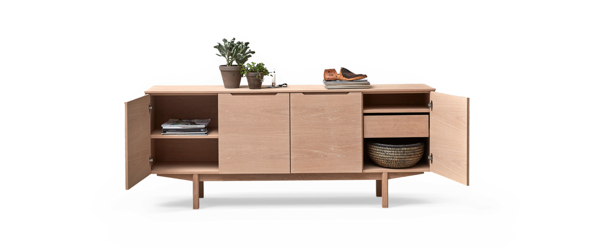 Ljus ekfärgat sideboard med öppna dörrar, hyllor och låda; böcker, växter, ljus och skor ovanpå samt flätad korg inuti.
