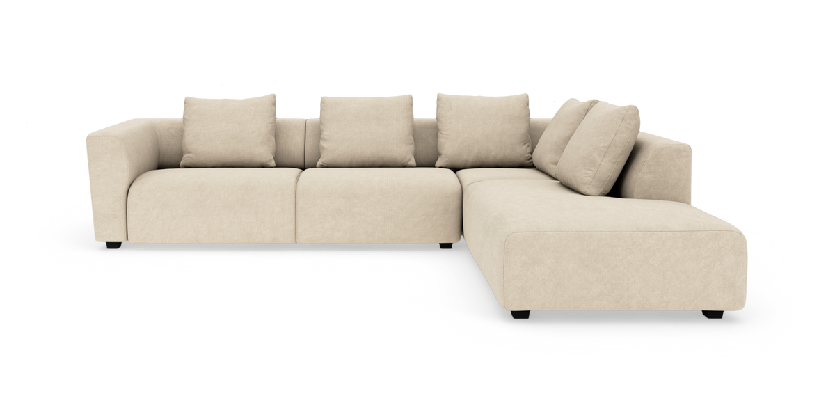 Beige L‑soffa med bred schäslong till höger och fyra lösa ryggkuddar, med rundade armstöd.