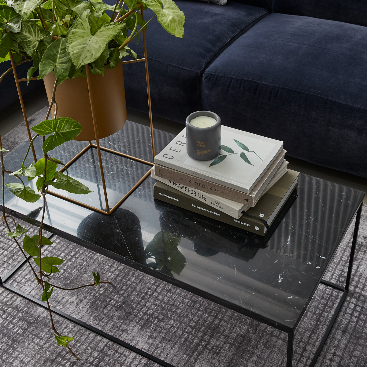 Svart marmorbord med grön krukväxt i guldfärgad ställning, stapel designböcker (Cereal, Kinfolk, A Frame for Life, The Design House Book, New Perspectives) och doftljus ”Silver Birch” på vardagsrumsbord.