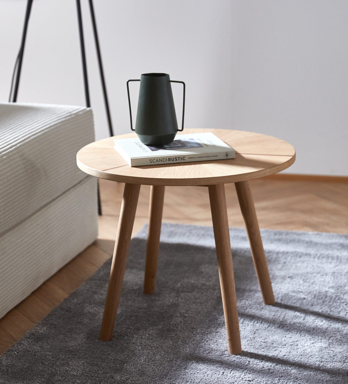 Runt sidobord i ek på grå matta bredvid soffa, med mörk kanna/vas ovanpå en bok med titeln SCANDIRUSTIC.