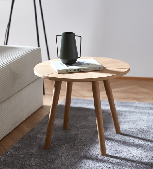Runt sidobord i ek på grå matta bredvid soffa, med mörk kanna/vas ovanpå en bok med titeln SCANDIRUSTIC.