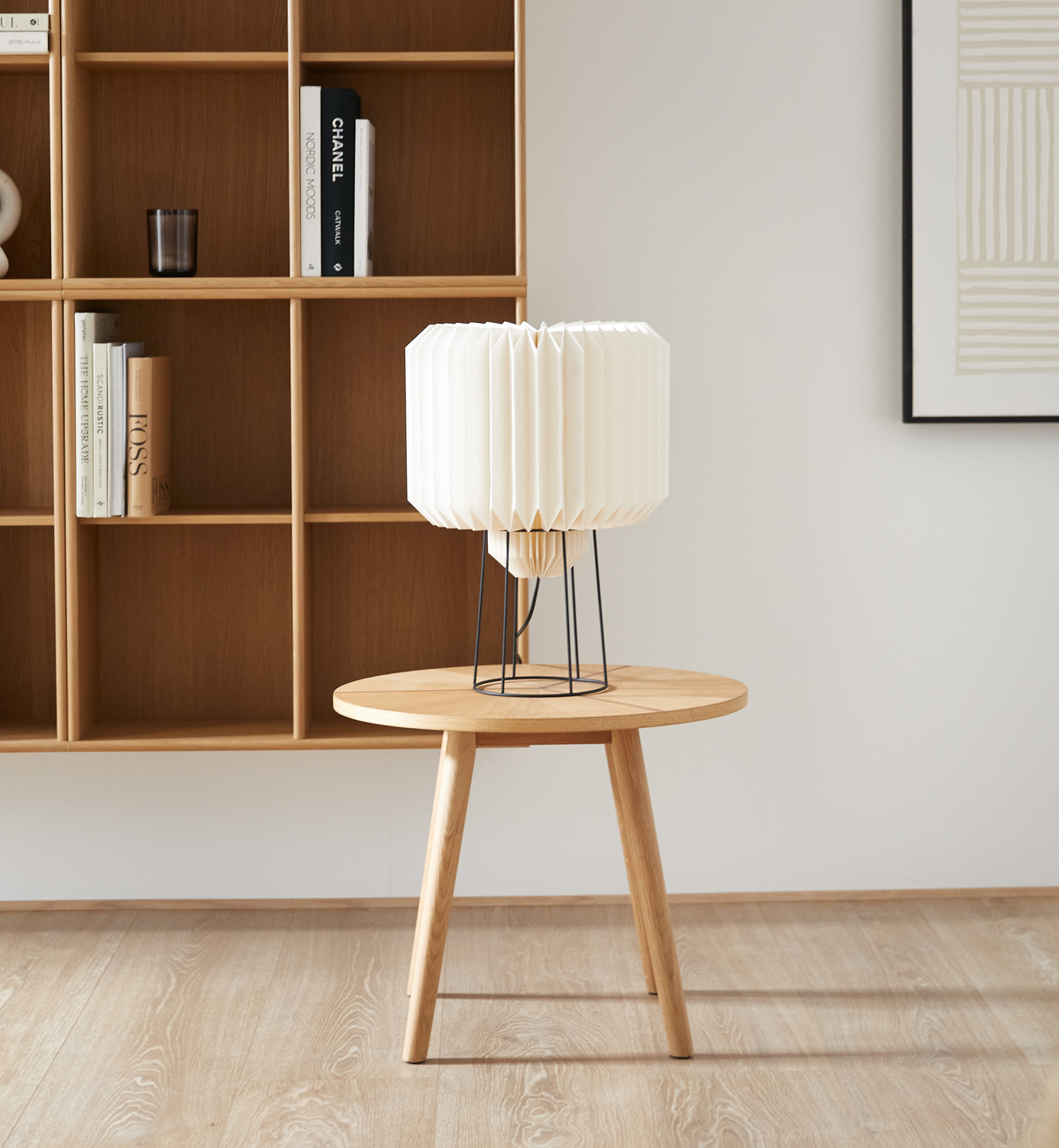 Ljust vardagsrum med runt sidobord i ek och vit plisserad bordslampa; i bakgrunden öppen hylla med böcker, bl.a. Chanel, och en minimalistisk tavla.