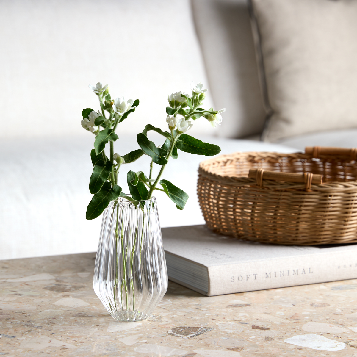 Räfflad glasvas med vita blommor på ett stenbord bredvid en flätad korg och en bok med titeln Soft Minimal i ett ljust vardagsrum.