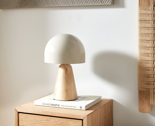 Beige bordslampa med träfot står på ”Scandi Rustic” av Rebecca Lawson & Reena Simon på ett skåp i ljust trä i ett ljust, luftigt rum.