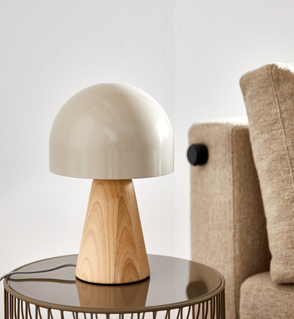 Bordslampa med krämfärgad kupol och träfot på ett runt sidobord bredvid en beige soffa i ett ljust vardagsrum.
