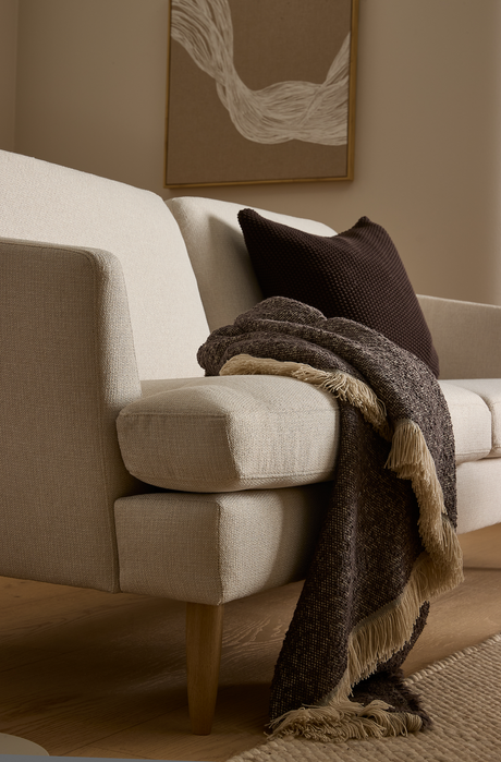Beige soffa med mörkbrun kudde och brun pläd med fransar över armstödet, träben, neutral matta och en abstrakt tavla på väggen.