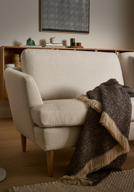 Beige tvåsitssoffa med träben och en brun fransad pläd över armstödet; i bakgrunden en skänk med böcker, ljus och korgar samt en abstrakt tavla.