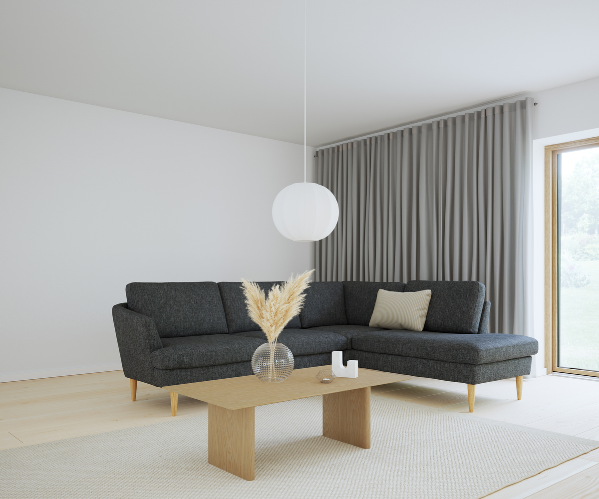 Ljust vardagsrum med mörkgrå hörnsoffa, träbord med glasvas med pampasgräs, beige matta, grå gardiner och vit rund pendellampa vid stort fönster.