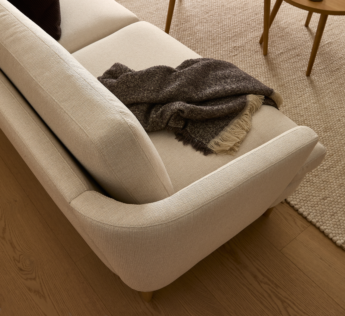 Beige soffa i vardagsrum med brun pläd med fransar, trägolv och ljus, texturerad matta.