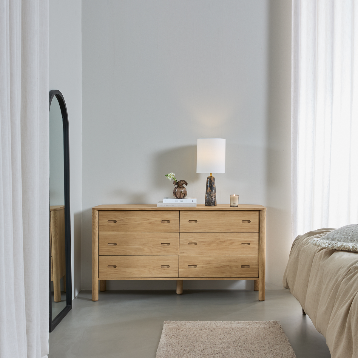 Ljust sovrum i neutrala toner med en ekbyrå med åtta lådor, bordslampa, spegel i golvhöjd, vas med grön kvist, ljus och bäddad säng.