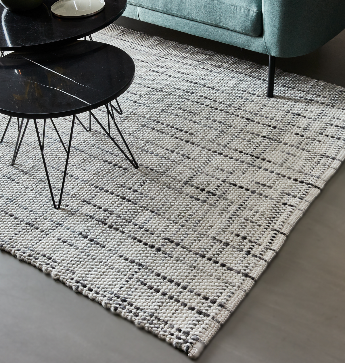 Ljusgrå vävd matta med diskret geometriskt mönster bredvid svarta runda soffbord och en turkos soffa i ett vardagsrum.