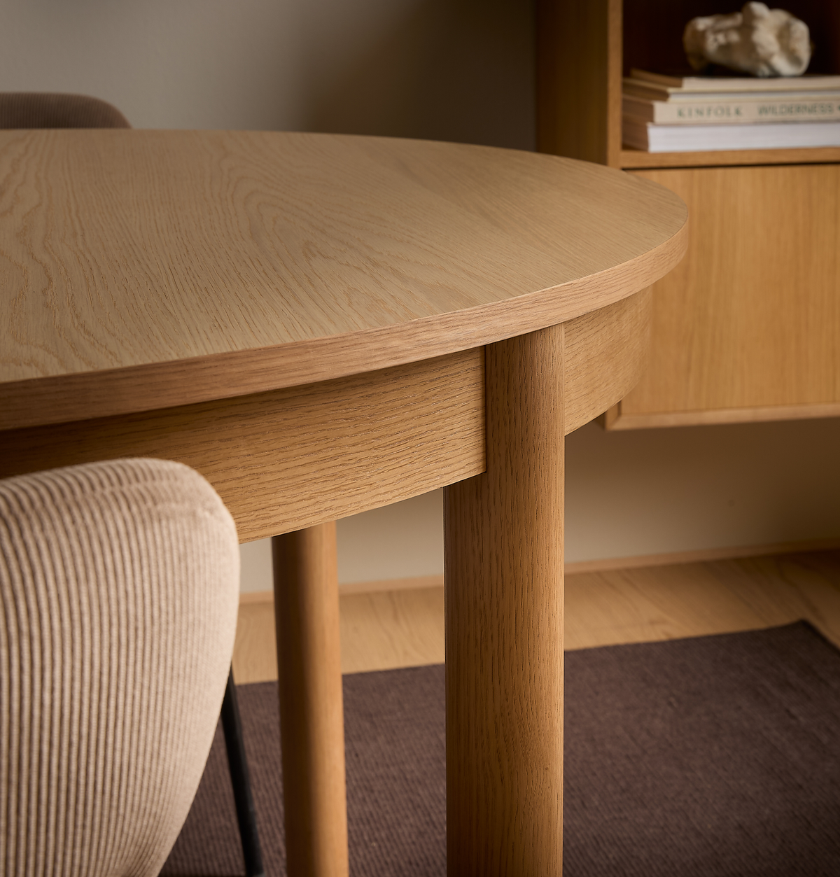 Närbild av ett rundat matbord i ljus ek med cylindriska ben och en beige ribbad stol; i bakgrunden en hylla med böcker – KINFOLK, WILDERNESS – och en dekorativ sten i en varm, minimalistisk interiör.