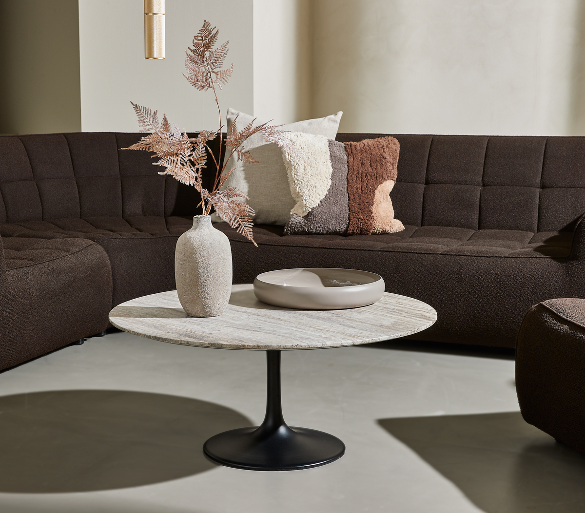 Modernt vardagsrum med mörk modulsoffa, runt soffbord med stenskiva och svart fot, beige vas med torkade blad och kuddar i bruna toner.