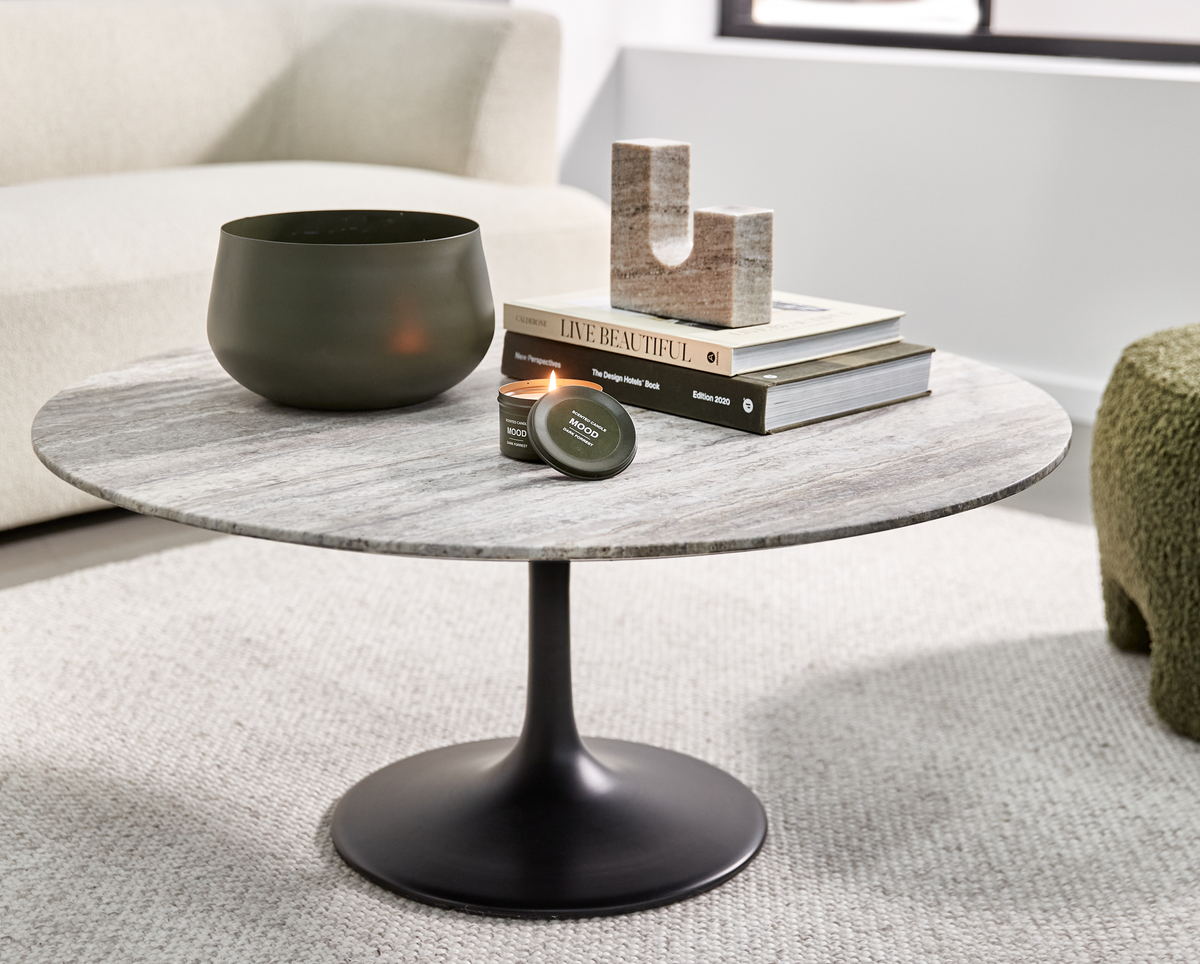 Runt soffbord med stenskiva och svart pelarfot; grön skål, tänd liten ljusburk märkt ”MOOD”, samt bokstapel med ”Live Beautiful” och ”The Design Hotels Book Edition 2020” och en U‑formad stenskulptur; ljus vardagsrumsmiljö.