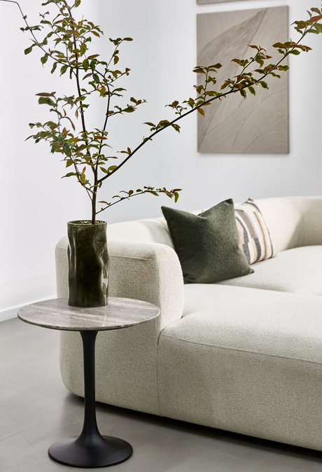 Ljus, minimalistisk vardagsrumsbild med ljus rundad soffa, grön kudde och en gren i mörkgrön vas på ett litet runt sidobord med stenbordsskiva; neutral väggkonst i bakgrunden.