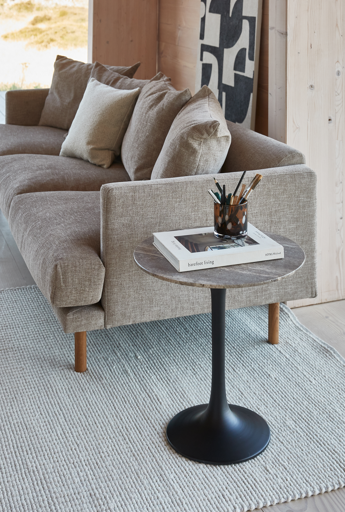 Beige soffa med runt sidobord i sten och svart fot, boken ”barefoot living” och pennor i kopp på en ljus matta i en skandinavisk vardagsrumsmiljö.