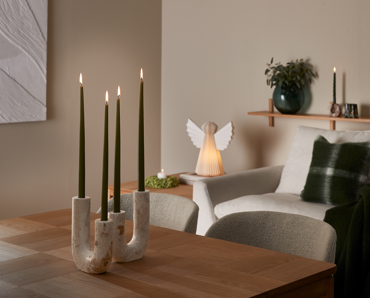 Fyra gröna kronljus i en skulptural ljusstake på ett träbord framför en ljus fåtölj och hylla med vas och tänd ängellampa i ett lugnt skandinaviskt vardagsrum.
