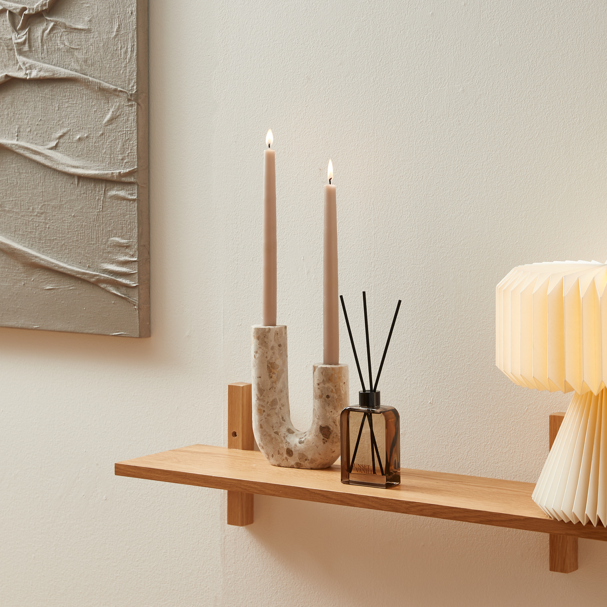 Trähylla med tända ljus i stenljusstake, doftpinnar och plisserad bordslampa mot ljus vägg.