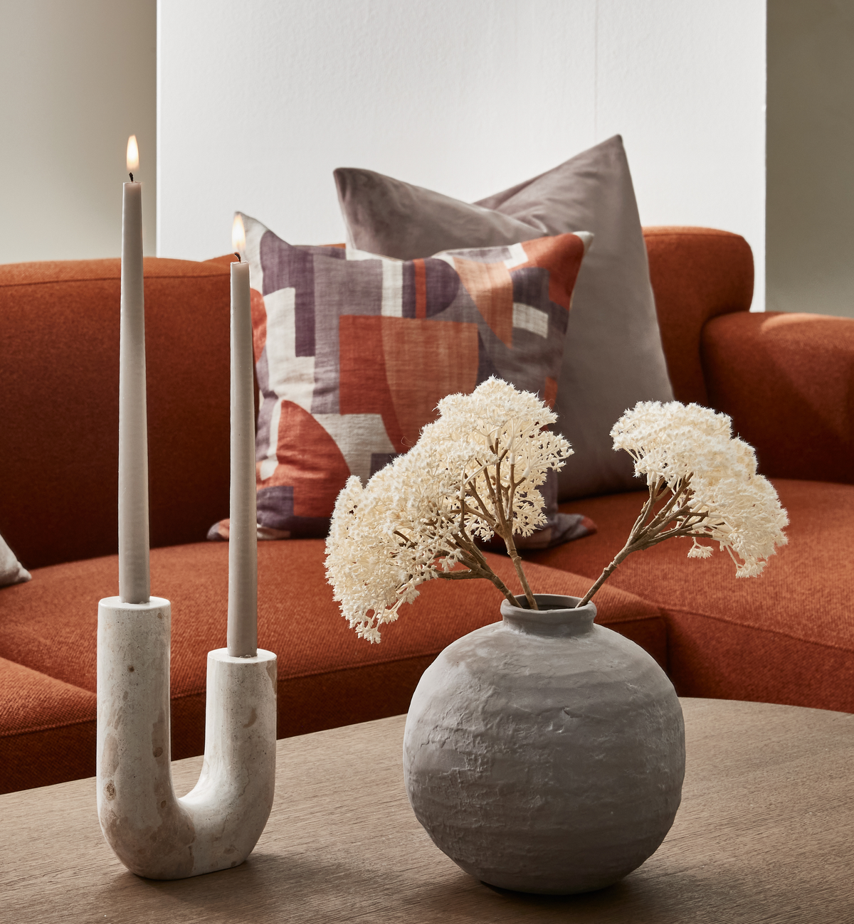 Grå keramikvas med vita torkade blommor och skulptural U‑formad ljusstake med två tända ljus på ett soffbord framför en roströd soffa med mönstrade kuddar.