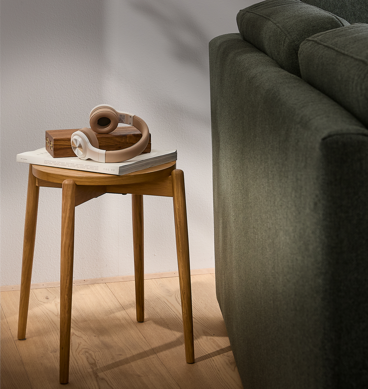 Litet runt sidobord i ljus ek bredvid en grön soffa, med beige hörlurar, en träask och en bok med titeln VOL VII i mjukt dagsljus.