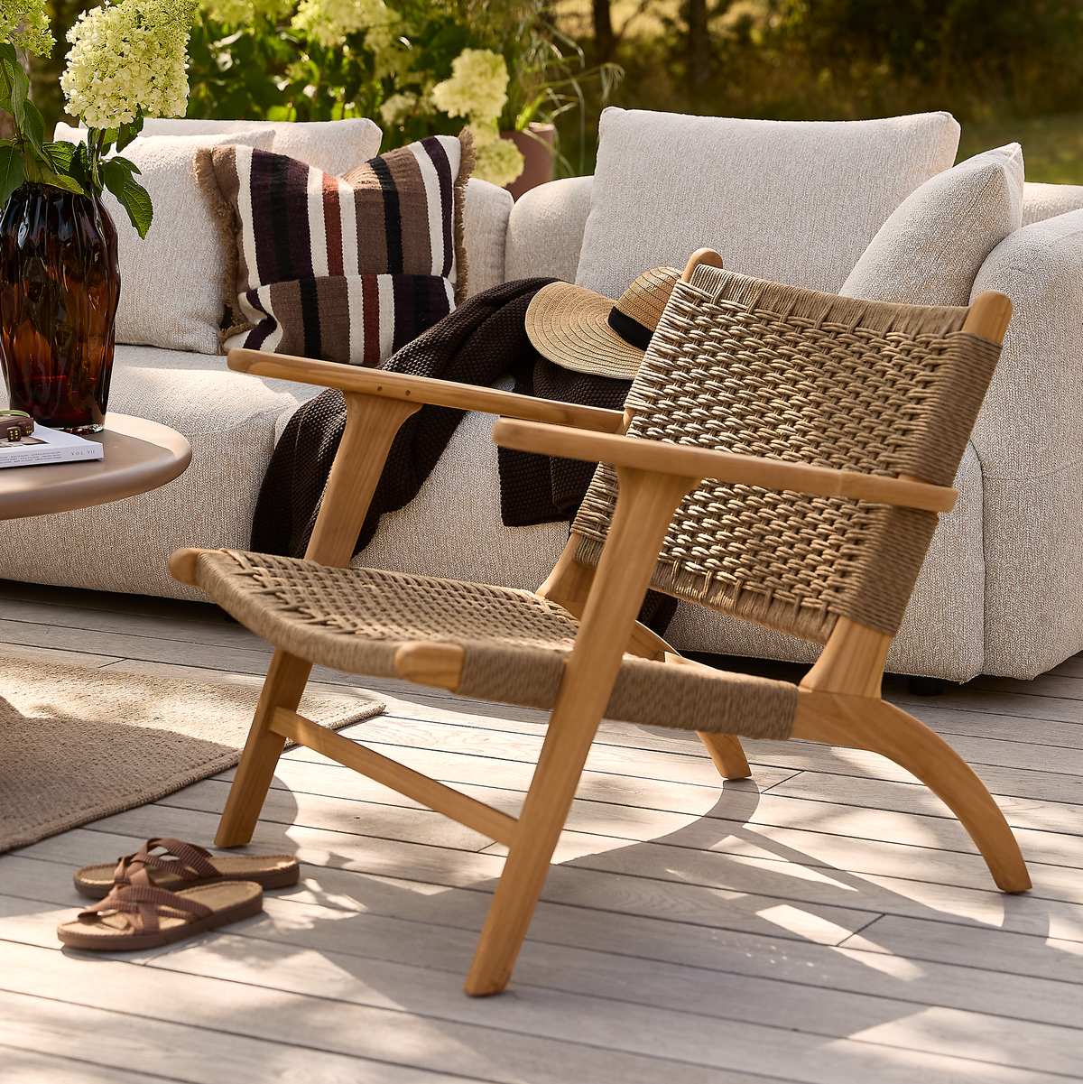 Låg loungefåtölj i trä med flätad sits på ljus trall, framför beige soffa med kuddar, filt och stråhatt; somrig uteplats med vas och sandaler i soligt ljus.