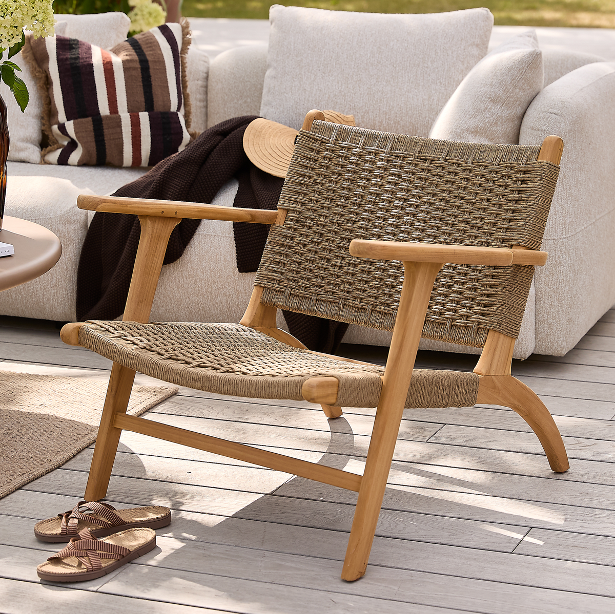 Låg loungefåtölj i trä med flätad sits och rygg på en solig terrass, framför en beige soffa med kuddar och filt; sandaler ligger på trädäcket.