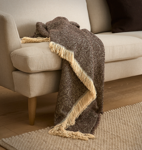 Brun melerad pläd med ljusa fransar draperad över en beige soffa, med ljus matta på trägolv.
