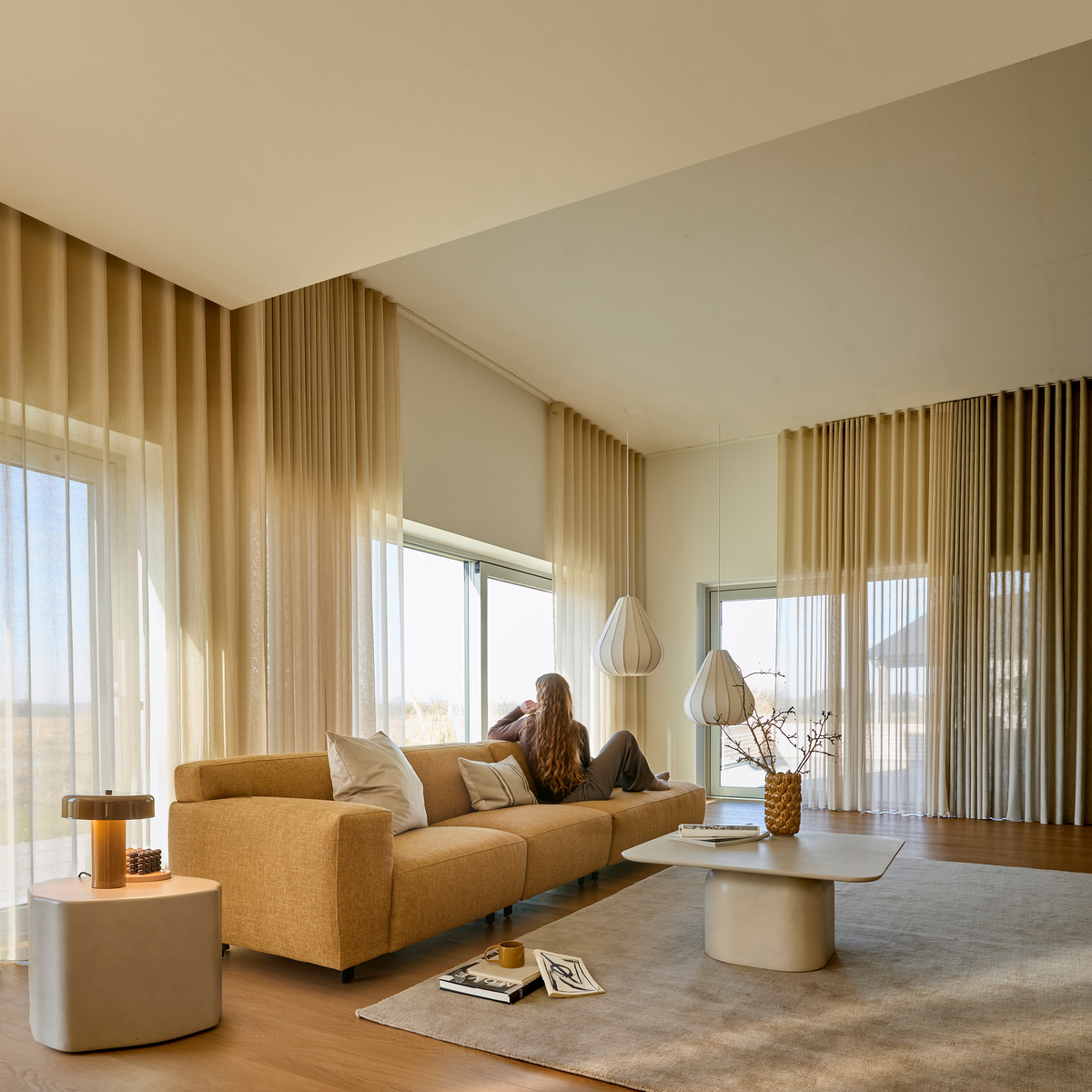 Ljust vardagsrum med beige soffa, stora fönster med skira gardiner, två vita pendellampor och lågt soffbord; en person sitter och blickar ut.