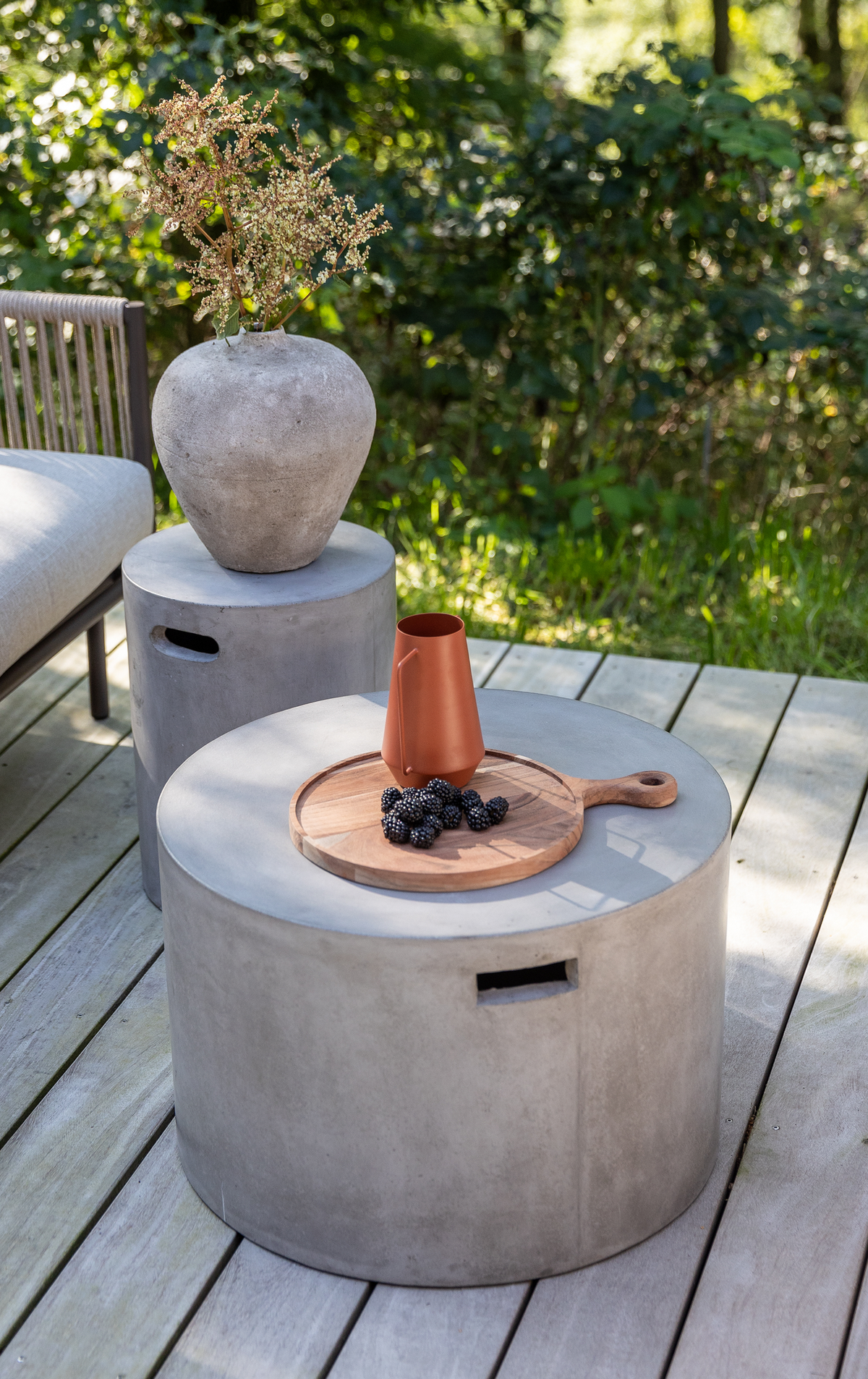 Runt betongbord på trädeck med träbricka, terrakottakanna och björnbär; bredvid ett cylindriskt sidobord med grå vas och torkade kvistar i grönskande utomhusmiljö.