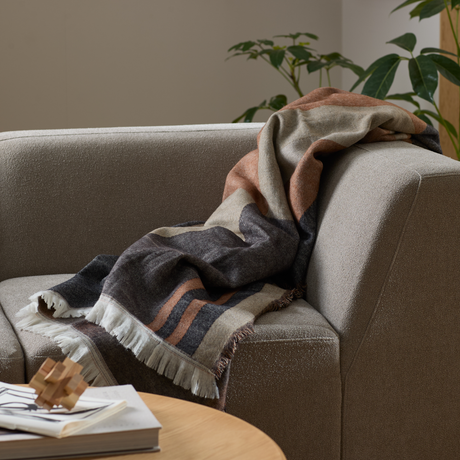 Randig pläd med fransar över armstödet på en beige soffa i ett vardagsrum med gröna krukväxter och ett träpussel på ett soffbord.