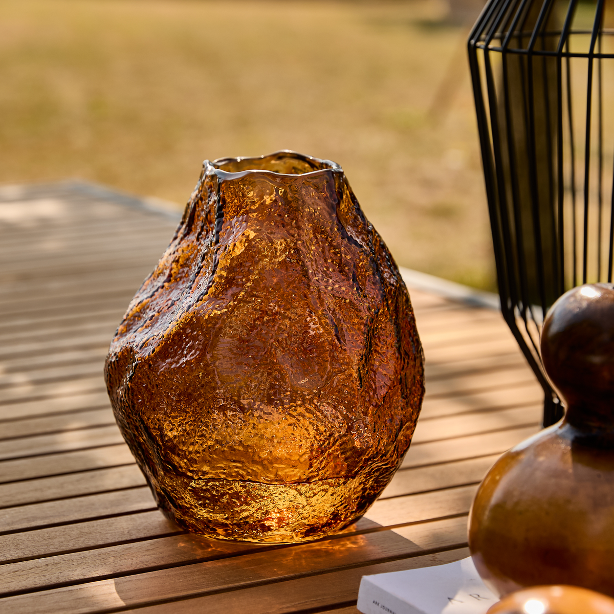 Bärnstensfärgad, strukturerad glasvas på ett träbord utomhus i varm eftermiddagssol.