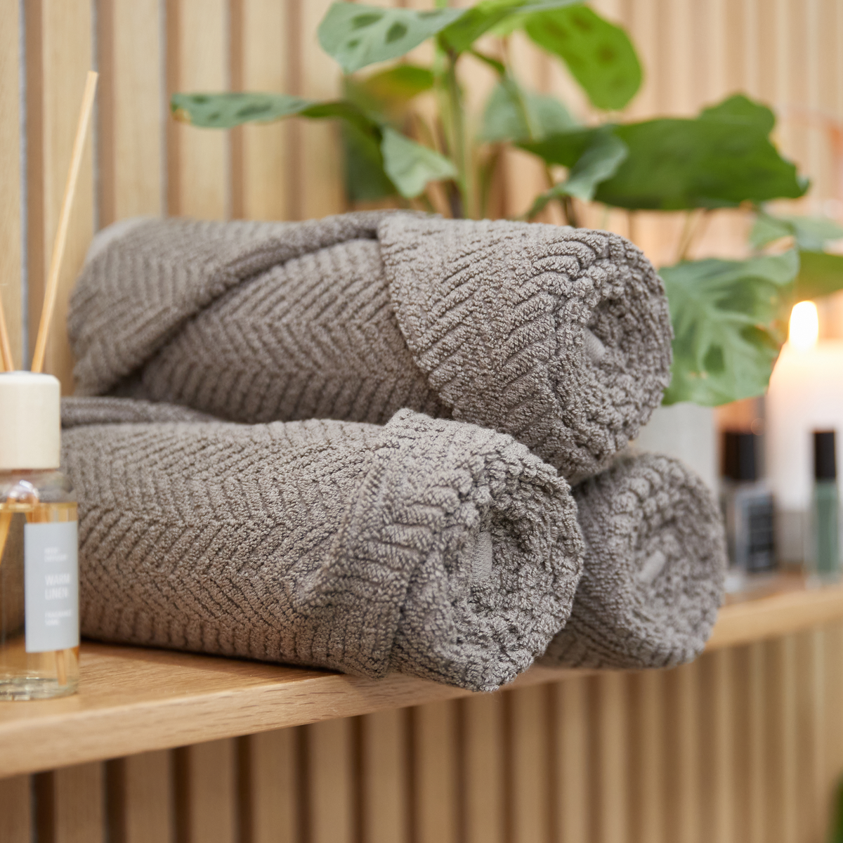 Tre rullade grå handdukar på en trähylla med grön växt, tända ljus och en doftspridare märkt ”WARM LINEN” i bakgrunden.