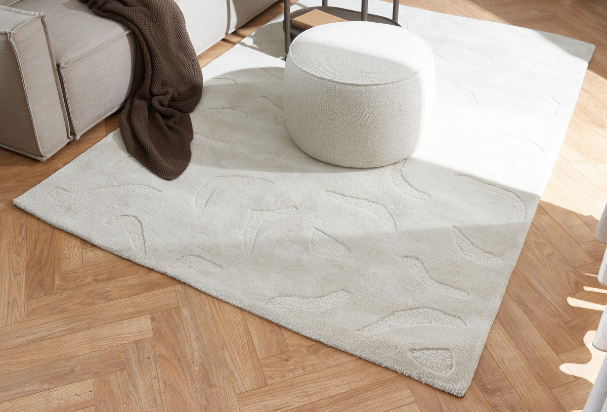 Ljusbeige matta med subtilt reliefmönster på fiskbensparkett, bredvid en vit puff och en beige soffa med brun pläd i dagsljus.