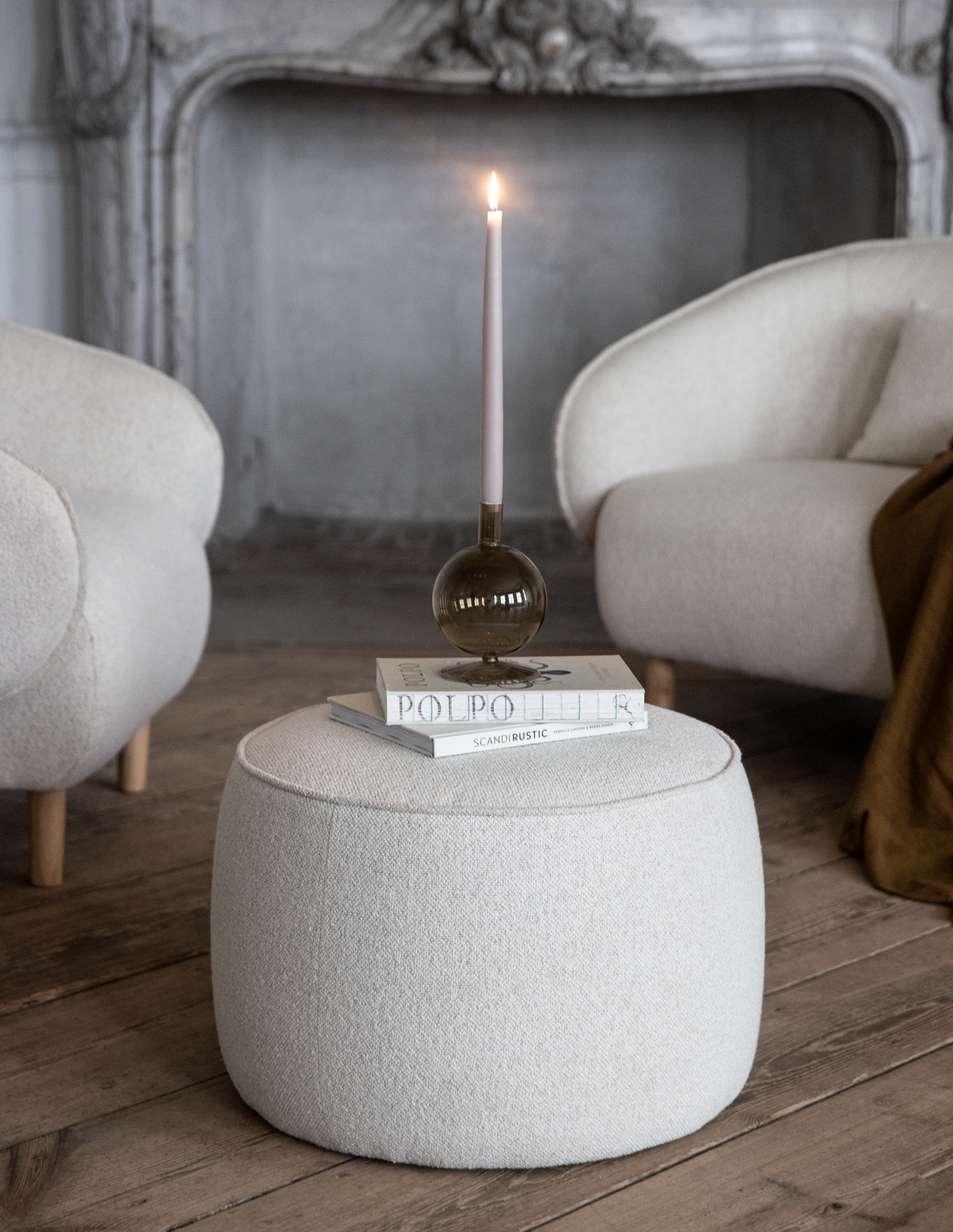 Beige puf i bouclé med böckerna ”POLPO” och ”SCANDI RUSTIC” ovanpå, toppad med en tänd smal ljus i rökfärgad glashållare mellan två ljusa fåtöljer vid en ornamenterad öppen spis.