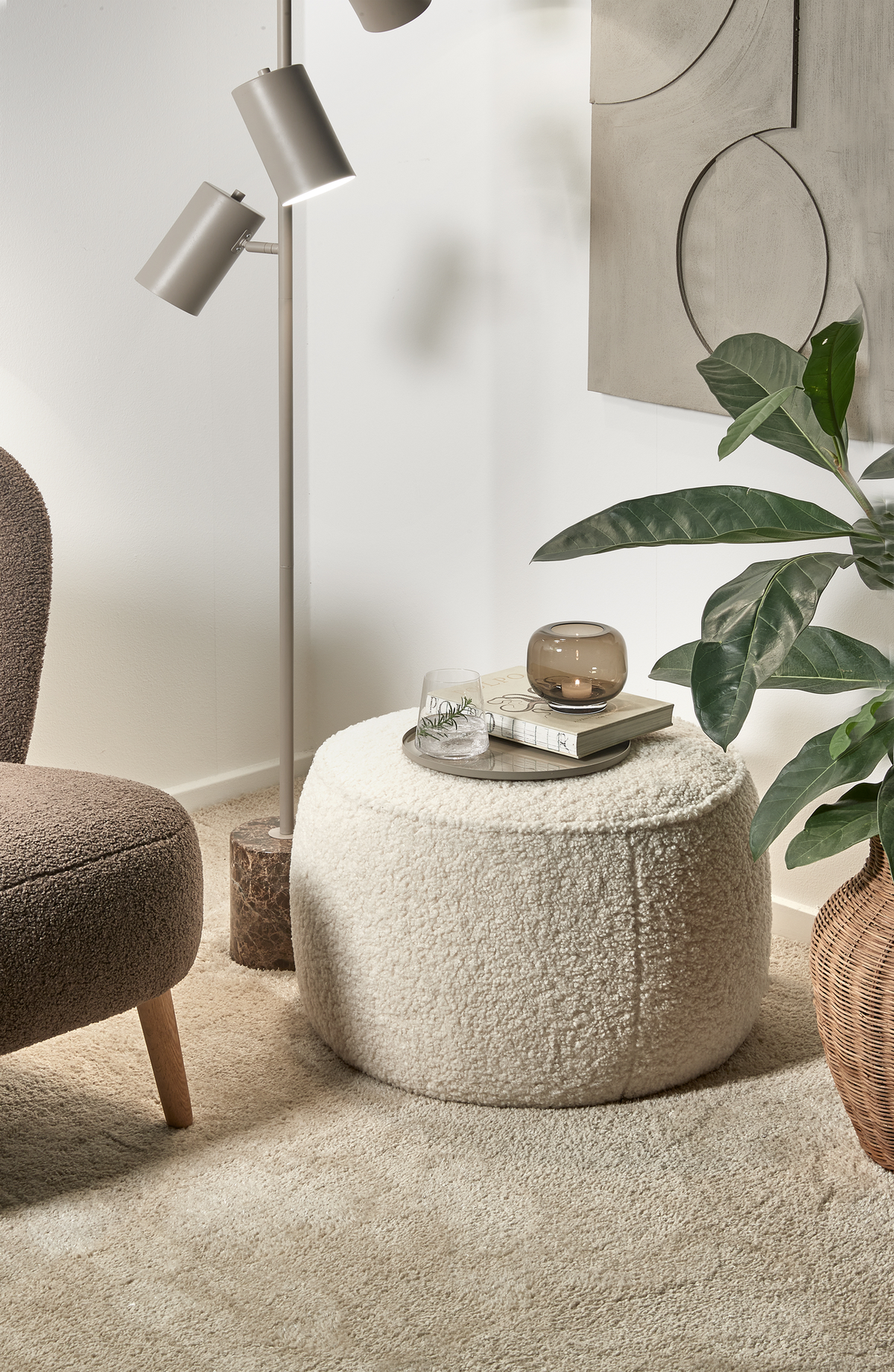 Mysig hörna i vardagsrum med beige bouclé‑puff som sidobord med bricka, glas och liten ljuslykta på, bredvid fåtölj, golvlampa, växt i korg och väggkonst i neutrala toner.