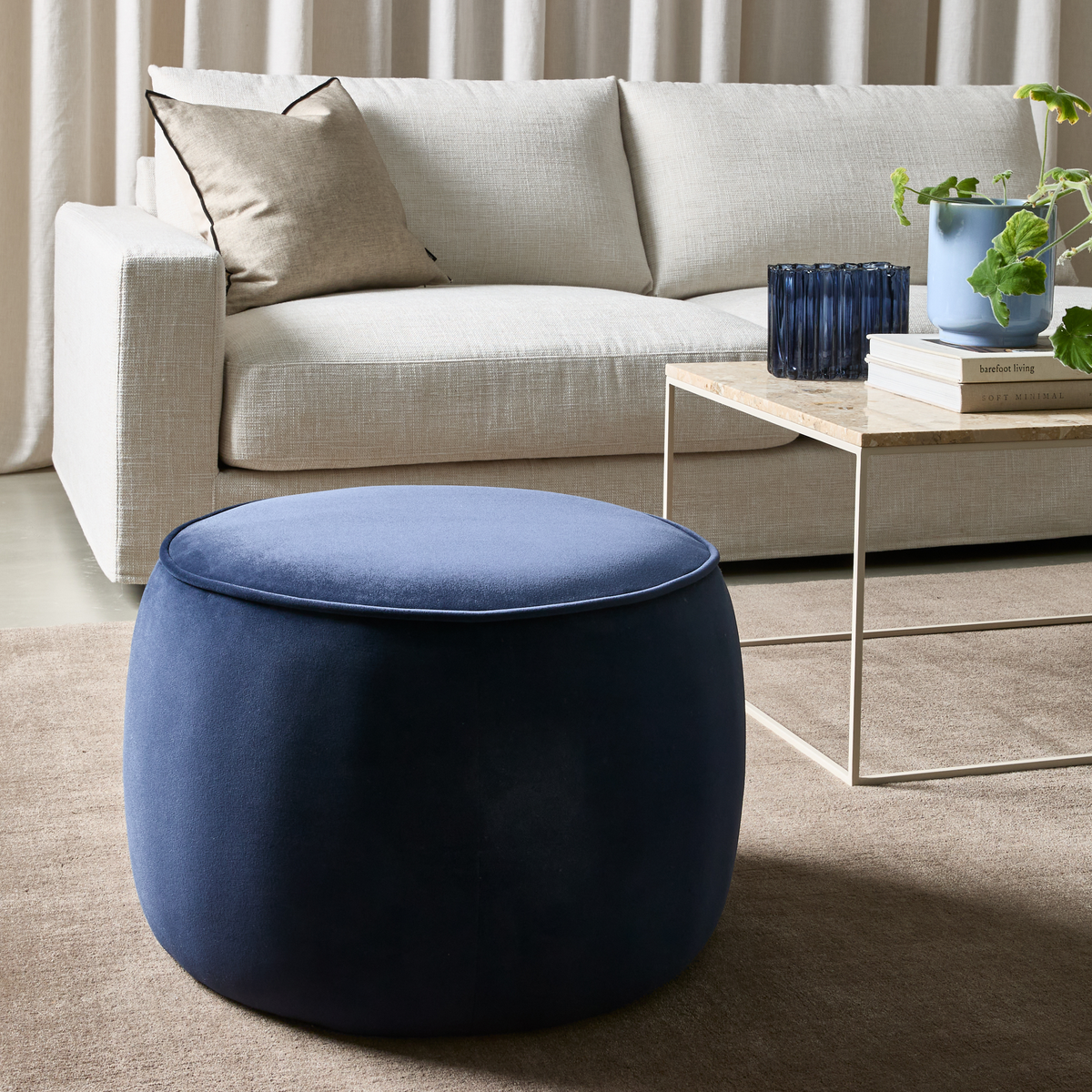 Beige soffa med kudde, ljust bord med bokhögar ”barefoot living” och ”Soft Minimal” bredvid blått glas och en blå kruka med växt; i förgrunden en rund mörkblå ottoman på beige matta.