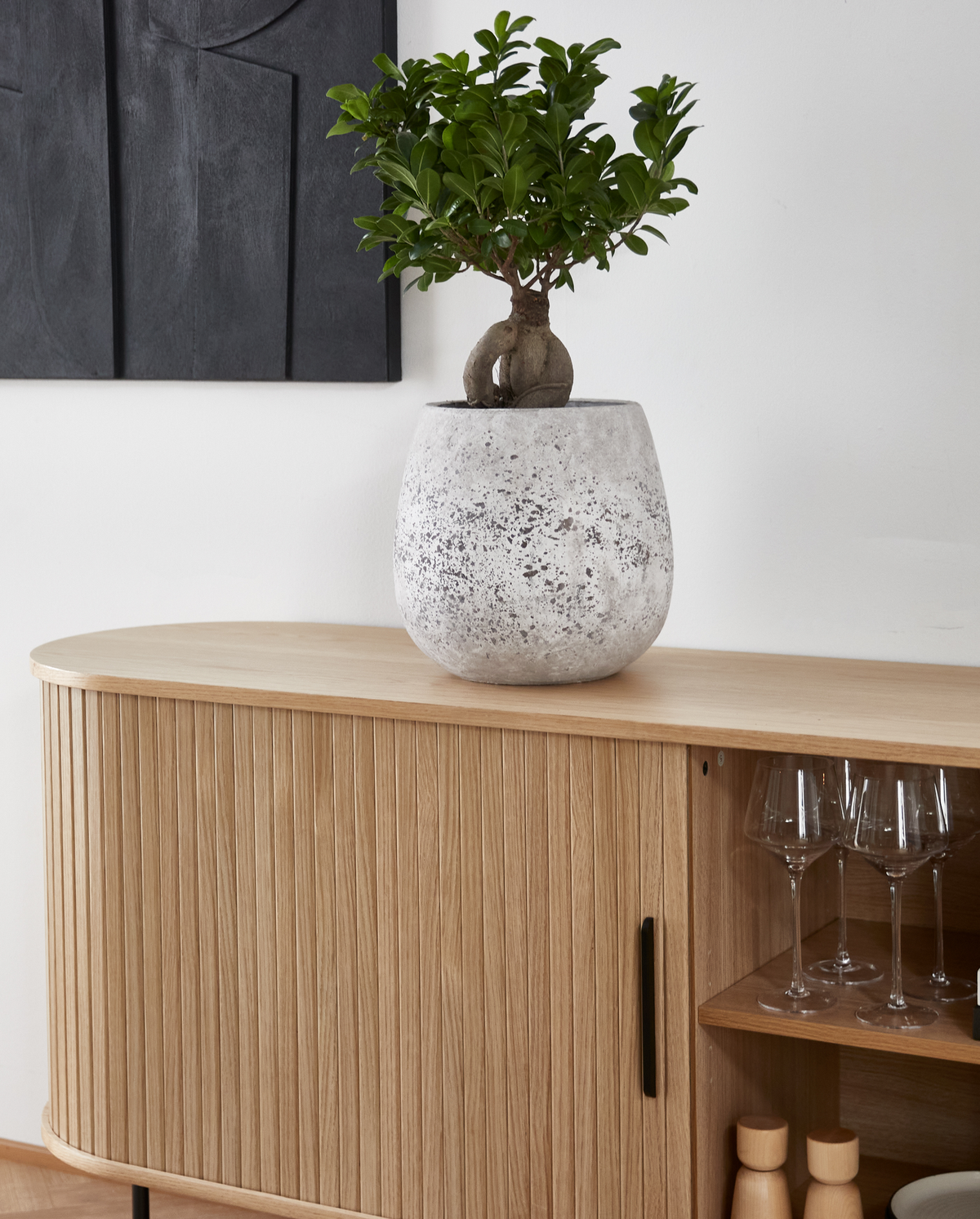 Liten grön krukväxt i grå stengodskruka på ribbad ek-sideboard med vinglas i öppen hylla.