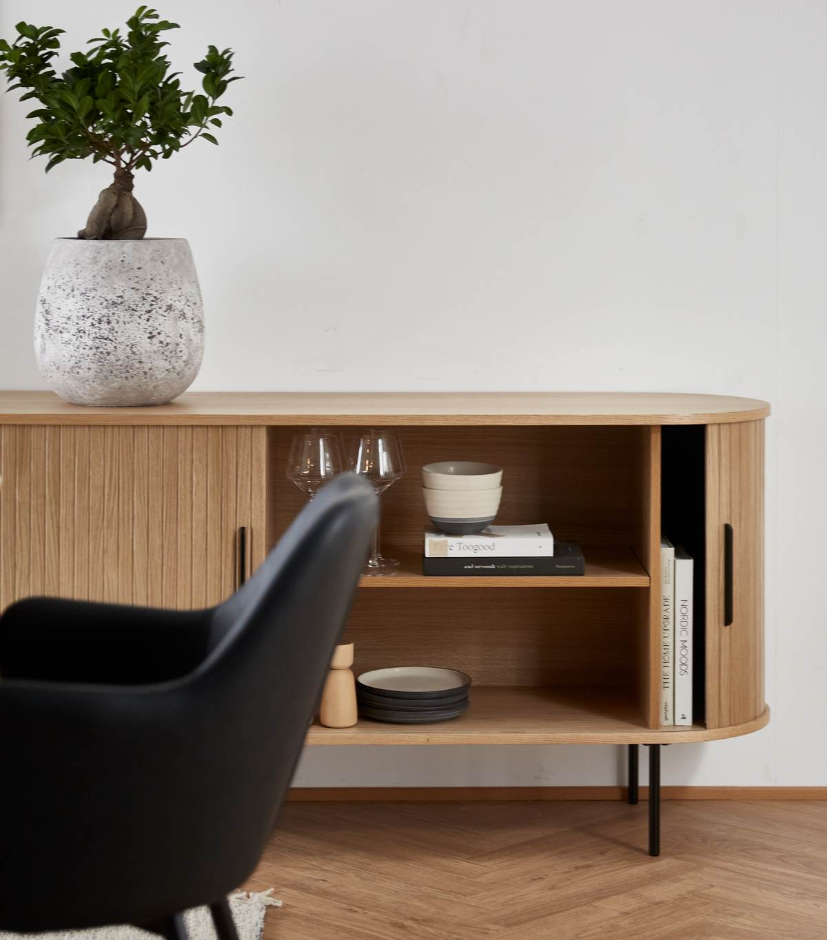 Ljus ek-skänk med öppna hyllor med skål, vinglas, staplade tallrikar, en liten kvarn och designböcker som ”Faye Toogood”; stor krukväxt ovanpå och en svart stol i förgrunden.