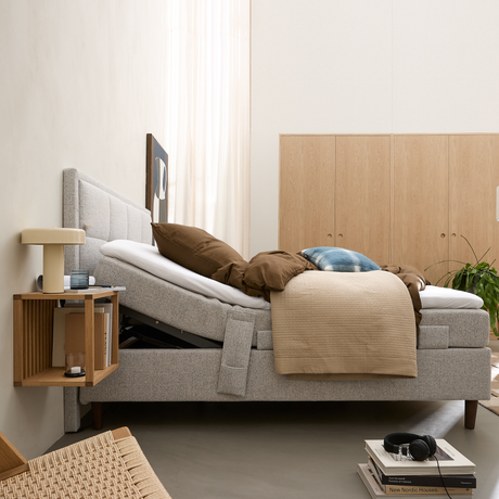 Ljust sovrum i skandinavisk stil med justerbar grå säng där huvudändan är uppfälld, beige och bruna sängkläder samt en blå kudde. Väggmonterat sängbord med liten bordslampa och glas. Ljus träförvaring och en grön växt i bakgrunden. På golvet ligger en hög böcker med hörlurar ovanpå; översta boken heter ”New Nordic Houses”.